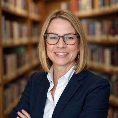 Headshot photo of Andrea Fleck-Nisbet in white shirt and dark blue jacket looking at the camera in front of shelves of books.